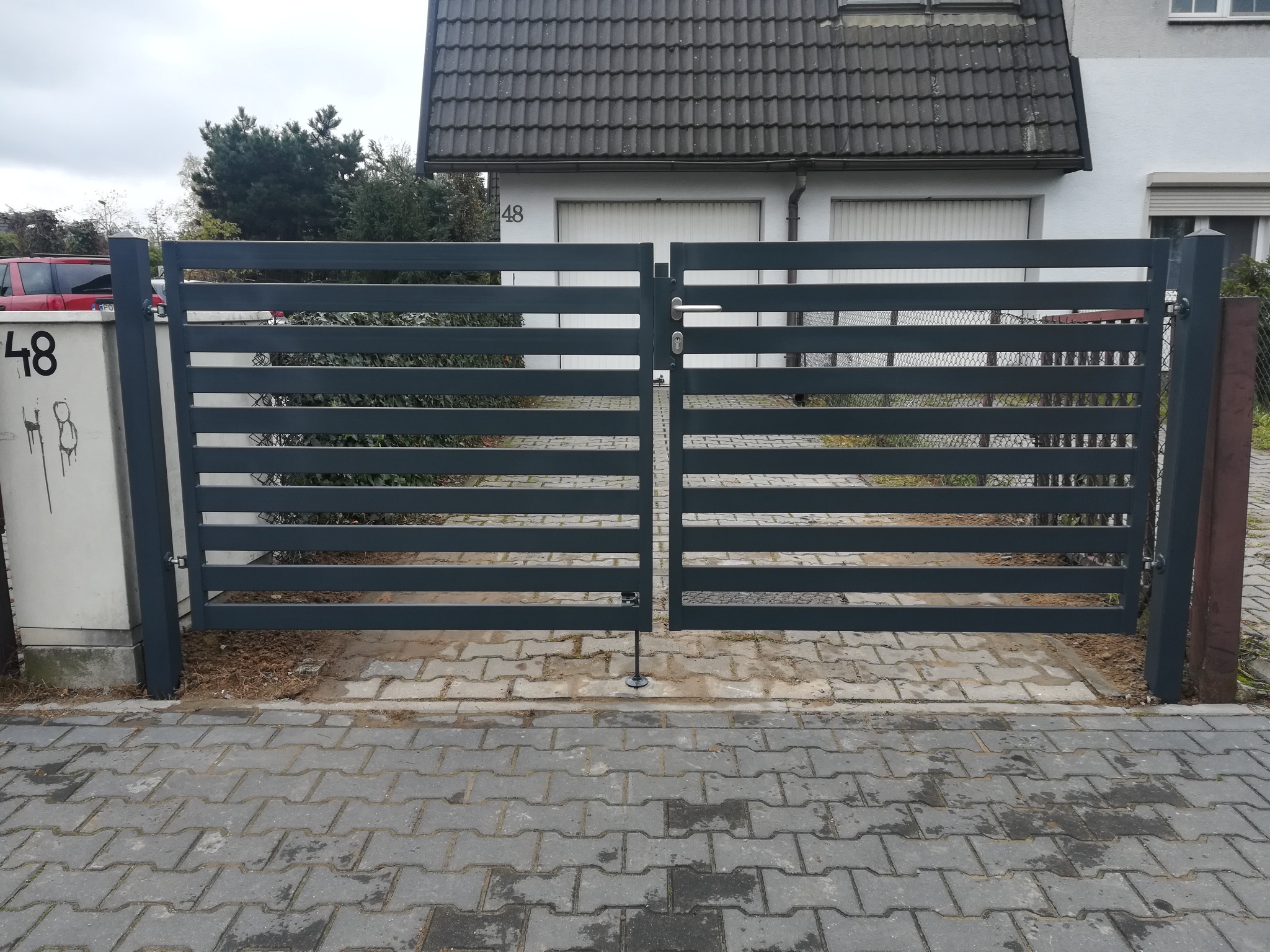Nowoczesna, pozioma brama wjazdowa w kolorze grafitowym, zamontowana na posesji z kostką brukową, widok na dom z numerem 48.