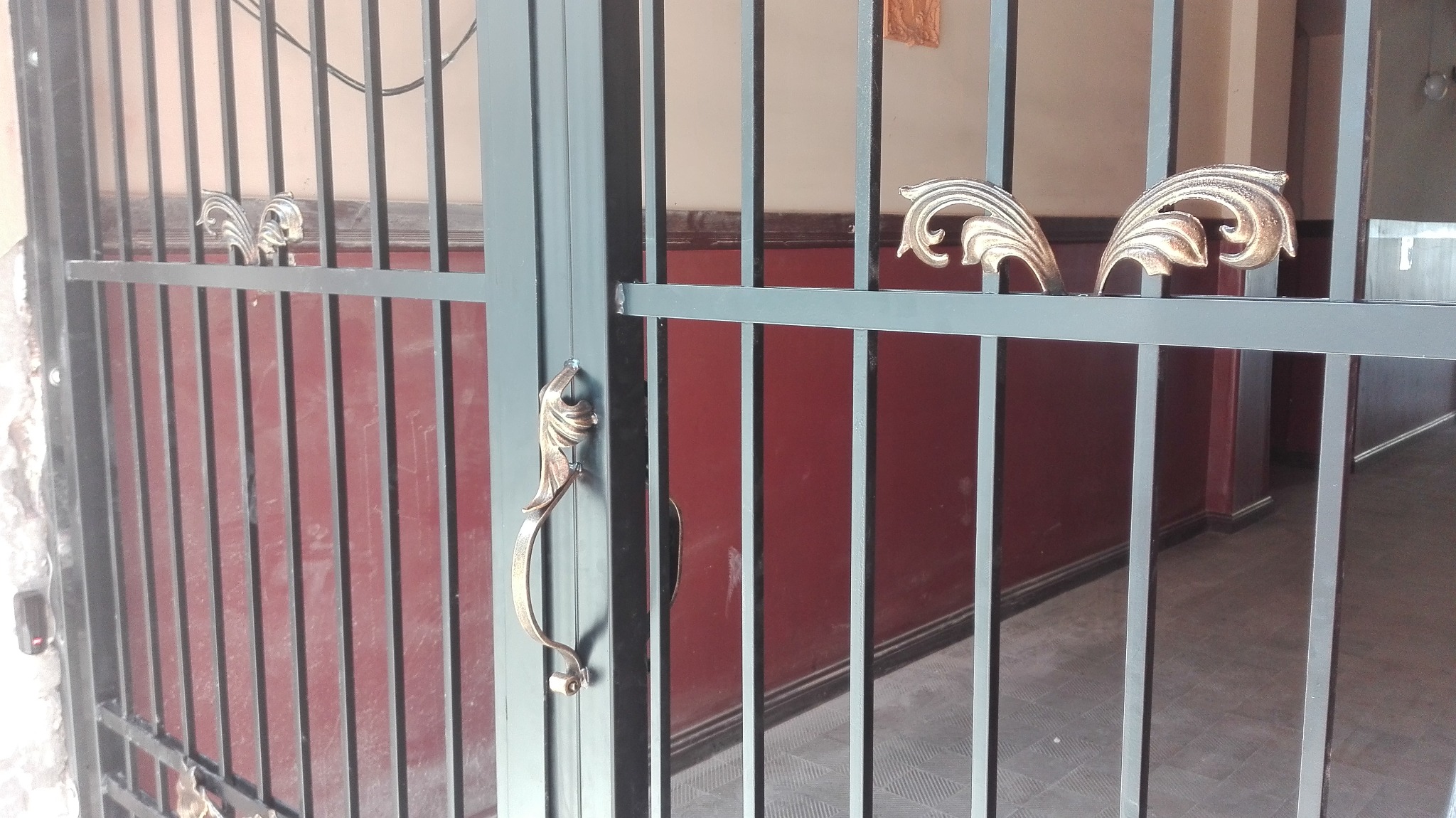 Szara metalowa krata z dekoracyjnymi elementami w kolorze złota, umieszczona wewnątrz budynku z czerwonymi ścianami.