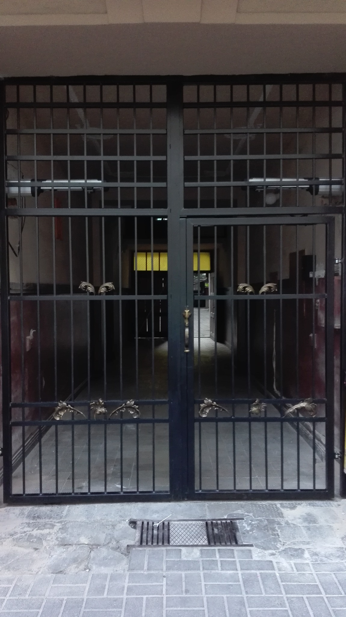 Metalowa brama z dekoracyjnymi elementami w kształcie liści i główek zwierząt, widok na korytarz z żółtym oświetleniem w głębi.