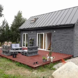 Szary, parterowy budynek z drewnianą elewacją i metalowym dachem, z tarasem z meblami z palet i małym psem rasy Jack Russell Terrier.