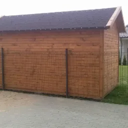 Drewniana altana z ciemnym dachem, widoczna z boku, z metalowym ogrodzeniem w tle. Konstrukcja stoi na kostce brukowej, obok trawnika i domu.