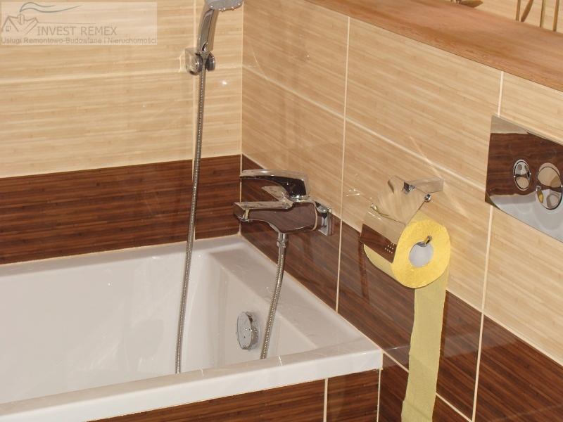 Wykończona łazienka z beżowo-brązowymi płytkami, białą wanną i chromowaną armaturą. Widoczny fragment toalety i żółty papier toaletowy.