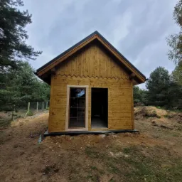 Surowa, drewniana konstrukcja domku z pojedynczym oknem i otwartymi drzwiami, na nierównym terenie z widocznymi śladami budowy, otoczona drzewami i pochmurnym niebem.