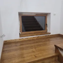 Wnętrze w trakcie wykańczania: drewniane schody z podestem, okno z widokiem na bieg schodów, białe ściany z widocznymi przewodami elektrycznymi.