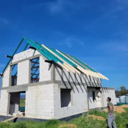 Niedokończony budynek z białych bloczków, z częściowo wykonaną konstrukcją dachu z zielonymi elementami, obserwowany przez mężczyznę w szarych spodniach na tle błękitnego nieba.