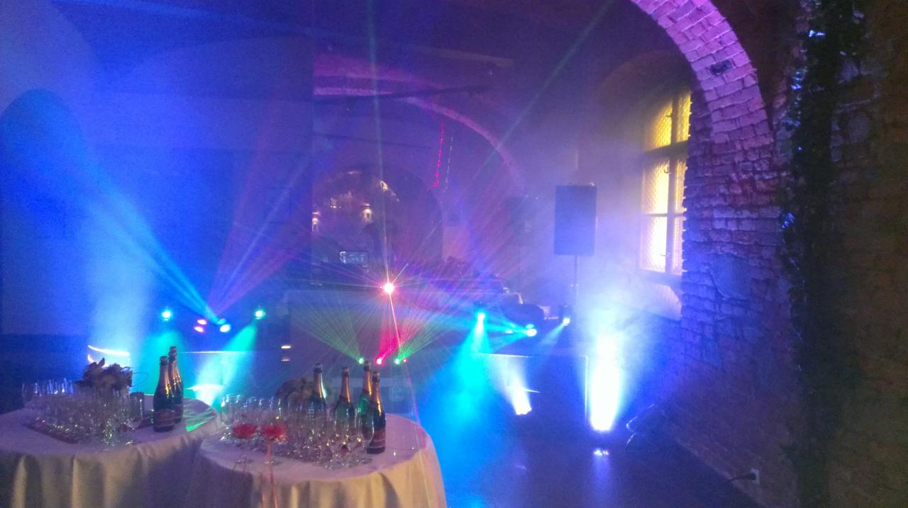 Uroczysta sala z nagłośnieniem i efektami świetlnymi: lasery, reflektory, stoliki z kieliszkami i butelkami, ceglany łuk w tle, tworzące imprezową atmosferę.