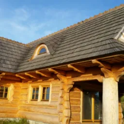 Zewnętrzny widok domu z bali z drewnianym dachem krytym gontem, okna z brązowymi ramami, kolumny podtrzymujące dach, błękitne niebo w tle.