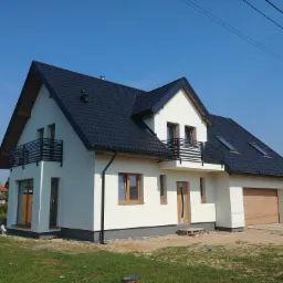 Nowoczesny dom jednorodzinny z białą elewacją, czarnym dachem i balkonem z metalową balustradą, widok z zewnątrz, jasne niebo.