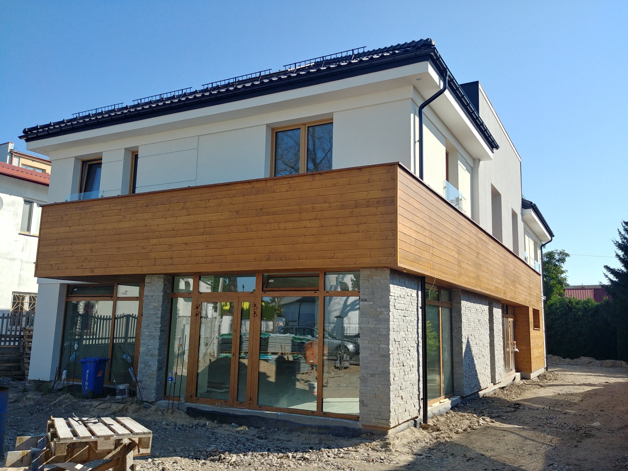 Nowoczesny dom dwukondygnacyjny z drewnianą okładziną na pierwszym piętrze i elementami kamiennej elewacji, w trakcie wykańczania z widocznymi materiałami budowlanymi i narzędziami na zewnątrz.