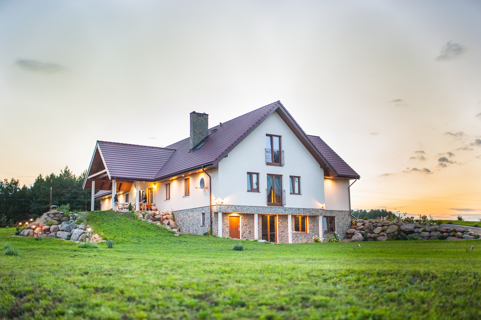 Duży, dwukondygnacyjny dom z białymi ścianami i ciemnoczerwonym dachem, otoczony zielonym trawnikiem i skalniakiem, oświetlony ciepłym światłem o zmierzchu.