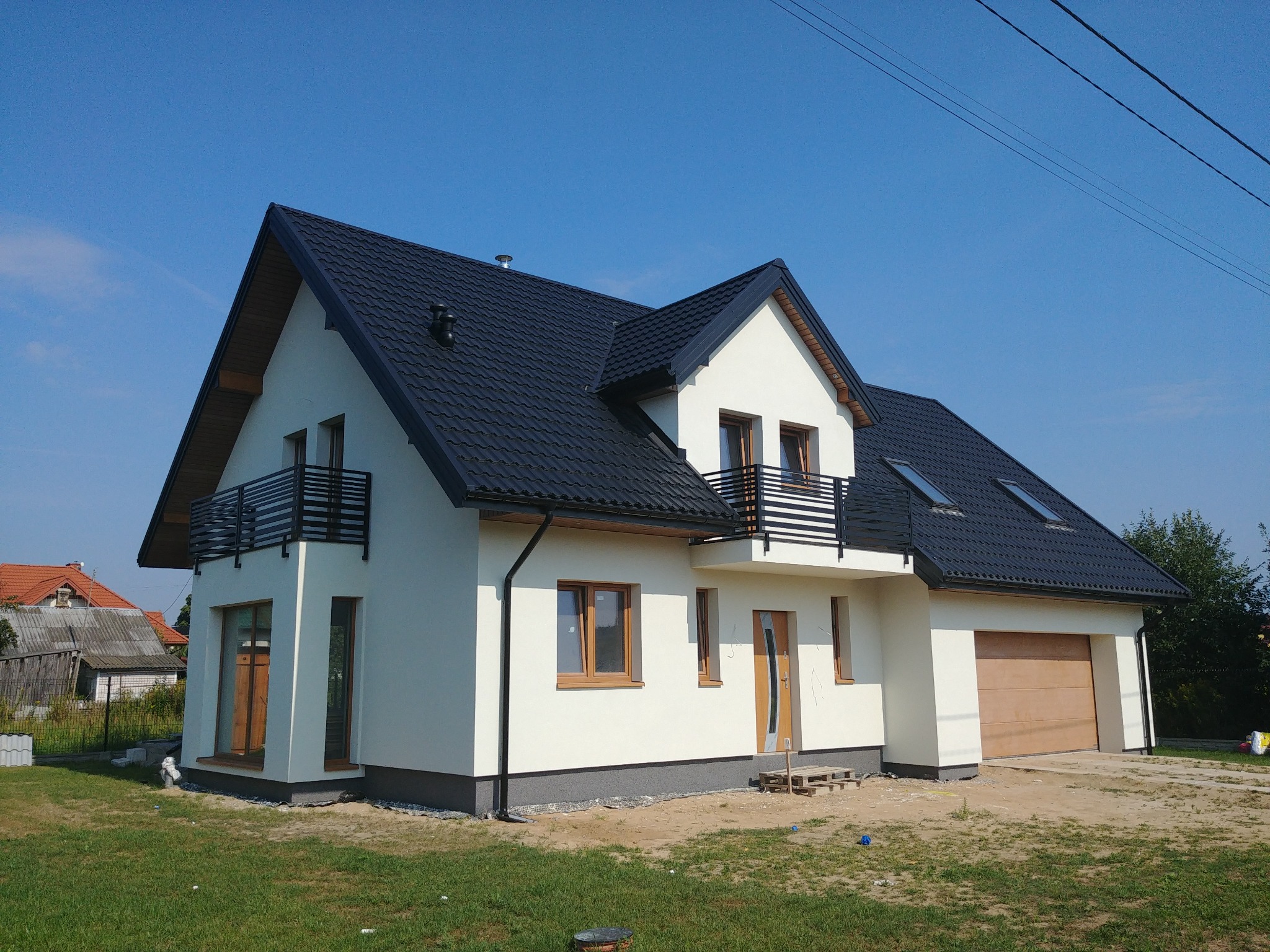 Nowoczesny dom jednorodzinny z białą elewacją, czarnym dachem i balkonem z metalową balustradą, widok z zewnątrz, jasne niebo.