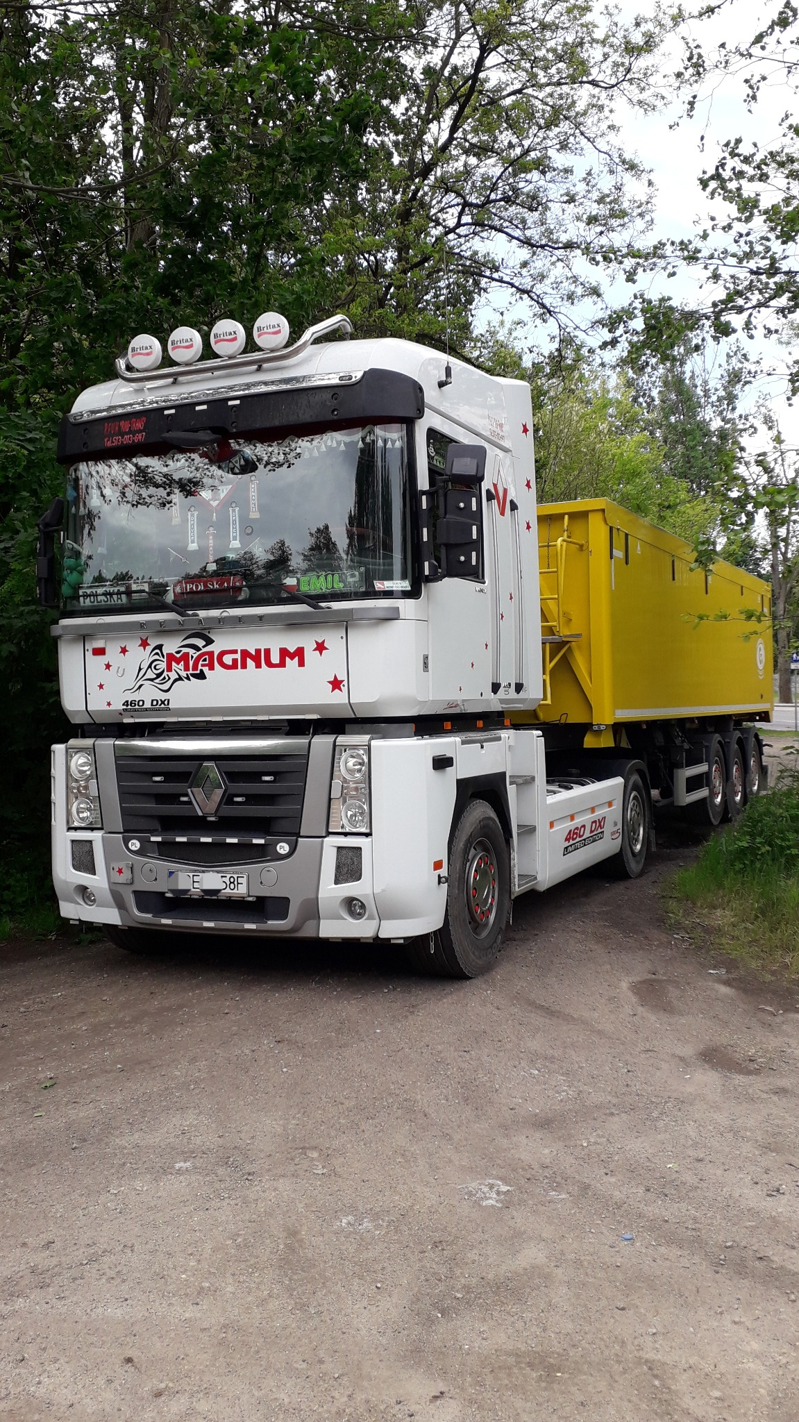 Biały Renault Magnum z żółtą naczepą kontenerową, ozdobiony gwiazdami i napisem Magnum, zaparkowany na żwirowej drodze, z widocznymi dodatkowymi lampami Britax na dachu kabiny.