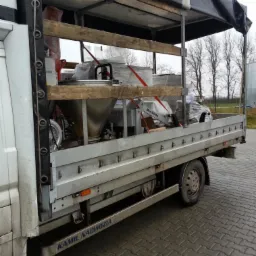 Częściowy widok z boku na bus z plandeką, przewożący zabezpieczone maszyny przemysłowe na drewnianych paletach, w tle drzewa i zbiornik.