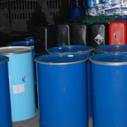 Kolorowe beczki i pojemniki z tworzyw sztucznych, niektóre z oznaczeniami ostrzegawczymi, ustawione w rzędach, prawdopodobnie gotowe do transportu lub magazynowania.