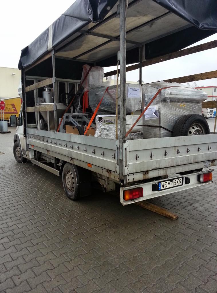 Otwarta ciężarówka z plandeką przewożąca maszyny owinięte folią i zabezpieczone pasami. Widoczny numer rejestracyjny WGM 12K9. Szara kostka brukowa.