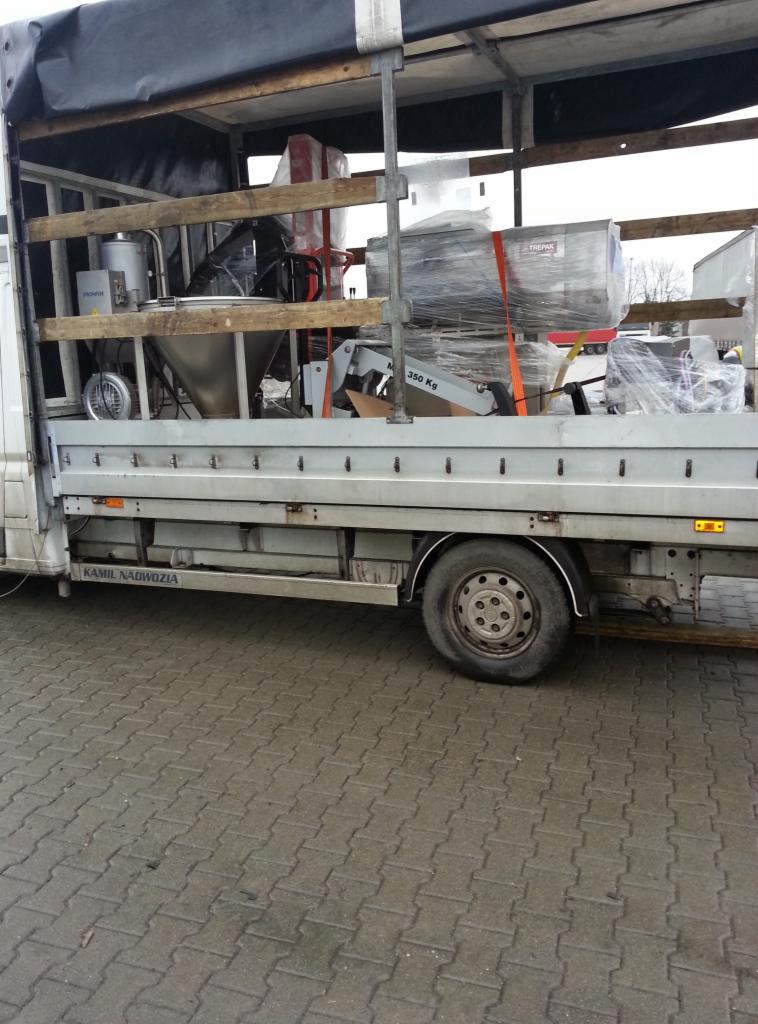 Bus z otwartą przestrzenią ładunkową przewozi zapakowane, zabezpieczone folią maszyny i urządzenia. Widoczne elementy konstrukcyjne pojazdu i opony.