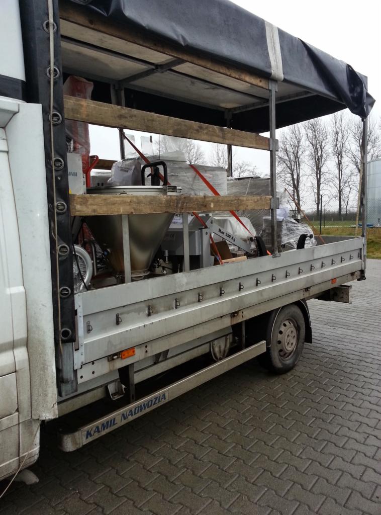 Częściowy widok z boku na bus z plandeką, przewożący zabezpieczone maszyny przemysłowe na drewnianych paletach, w tle drzewa i zbiornik.