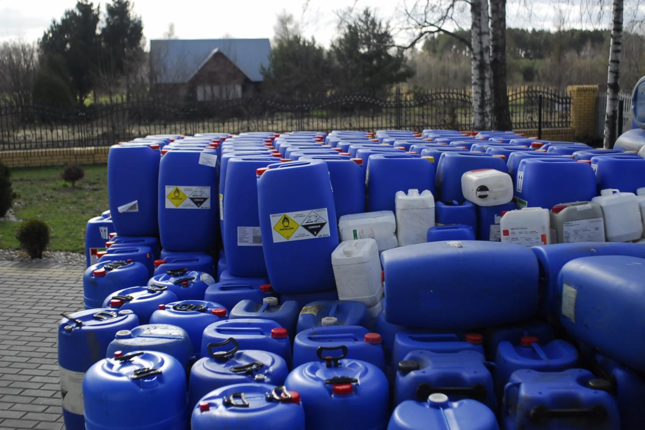 Stos niebieskich i białych pojemników z etykietami ostrzegawczymi na zewnątrz, z widocznym budynkiem w tle.