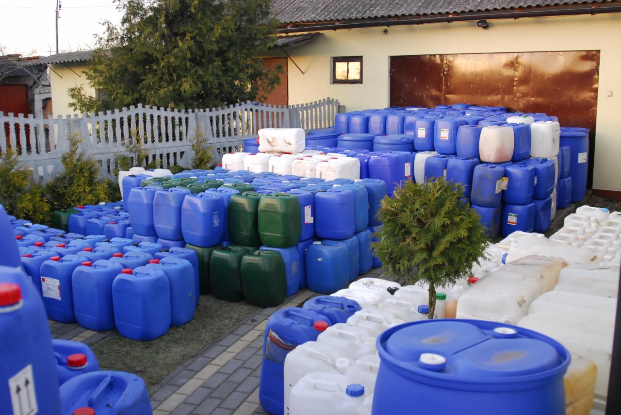 Magazyn pełen niebieskich, białych i zielonych pojemników plastikowych, różnej wielkości, ułożonych na zewnątrz budynku z brązową bramą garażową i białym płotem.