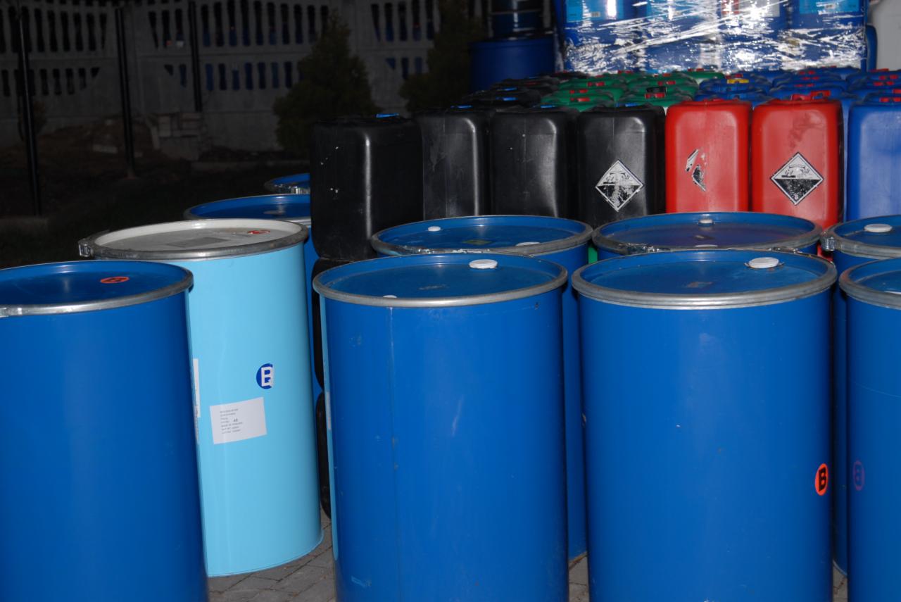 Kolorowe beczki i pojemniki z tworzyw sztucznych, niektóre z oznaczeniami ostrzegawczymi, ustawione w rzędach, prawdopodobnie gotowe do transportu lub magazynowania.
