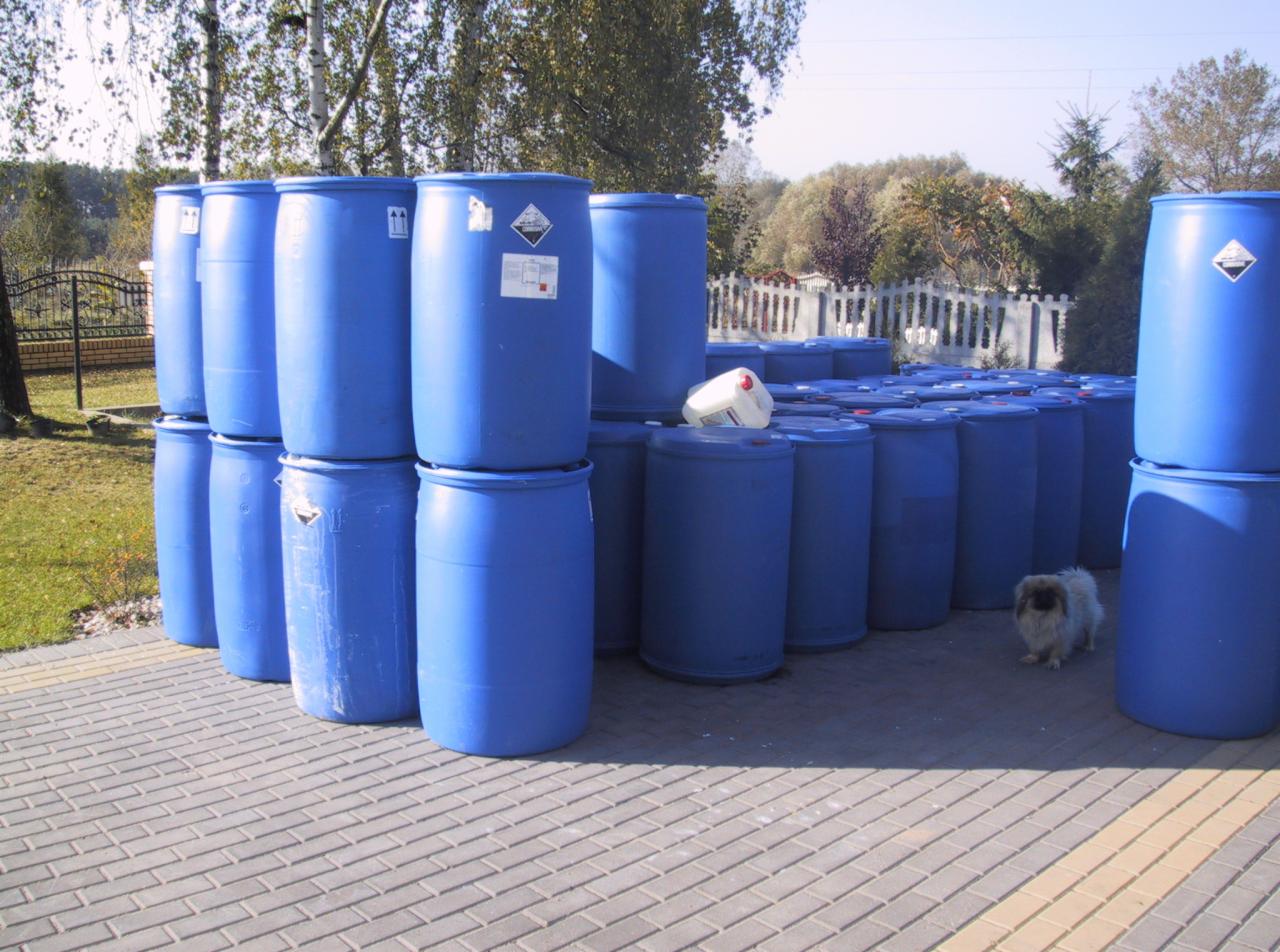 Stos niebieskich beczek z etykietami ostrzegawczymi na kostce brukowej, w tle drzewa i krzewy. Pies rasy Shih Tzu stoi obok. Jedna beczka biała.