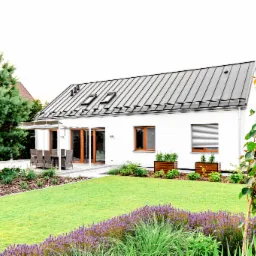 Nowoczesny dom jednorodzinny z białymi ścianami i ciemnym, metalowym dachem, otoczony zadbanym ogrodem z zielonym trawnikiem i rabatami kwiatowymi, widoczne drewniane okna i drzwi tarasowe.