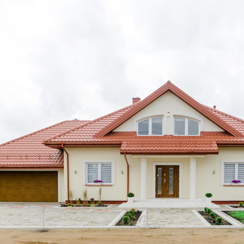 Gotowy dom jednorodzinny z beżową elewacją, czerwoną dachówką, drewnianymi drzwiami wejściowymi, garażem i zagospodarowanym terenem przed budynkiem.