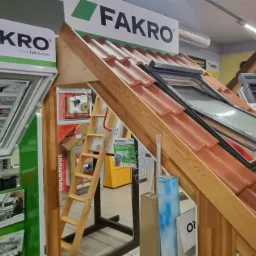 Ekspozycja okien dachowych Fakro i Velux z drewnianą konstrukcją imitującą dach, prezentowana w sklepie z materiałami budowlanymi. Widoczna drabina i reklamy produktów.