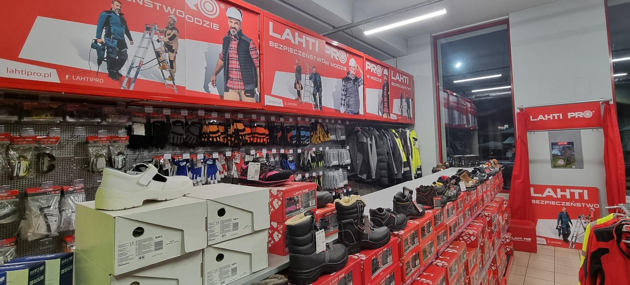 Wnętrze sklepu z odzieżą i obuwiem roboczym Lahti Pro, prezentujące różnorodny asortyment, w tym białe buty robocze na pudełkach, rękawice ochronne i kurtki, z reklamami marki na górze.
