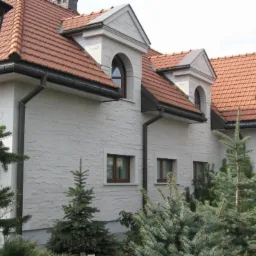 Elewacja domu jednorodzinnego z jasną, strukturalną ścianą, czerwoną dachówką i oknami w brązowych ramach, oświetlona przez czarne, ozdobne latarnie parkowe.