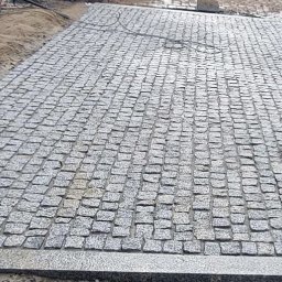 MOFFIJ Bruk - Świeżo ułożona, szara kostka granitowa tworzy równą nawierzchnię. Widoczne worki z materiałem budowlanym i sprzęt w tle. Krawężnik oddziela kostkę od jezdni.
