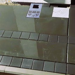 MOFFIJ Bruk - Świeżo ułożona, ciemna kostka brukowa z odpływem liniowym i studzienką rewizyjną. Widoczne obrzeża i fuga. Całość sprawia wrażenie mokrej, być może po myciu.