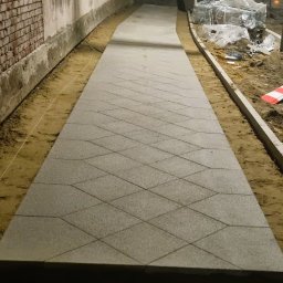 MOFFIJ Bruk - Ścieżka z szarej kostki o wzorze rombów, otoczona piaskiem i ceglanym murem. Widoczne materiały budowlane w tle. Ujęcie z perspektywy.