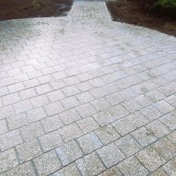 MOFFIJ Bruk - Ścieżka z jasnoszarej kostki granitowej ułożona łukiem, otoczona ziemią i zielenią. Widoczny fragment ścieżki z kostki w odcieniach brązu.