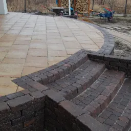 Ułożona kostka brukowa w odcieniach beżu i brązu tworzy schody i taras. W tle widoczne narzędzia ogrodowe i drewno opałowe, co sugeruje prace wykończeniowe.