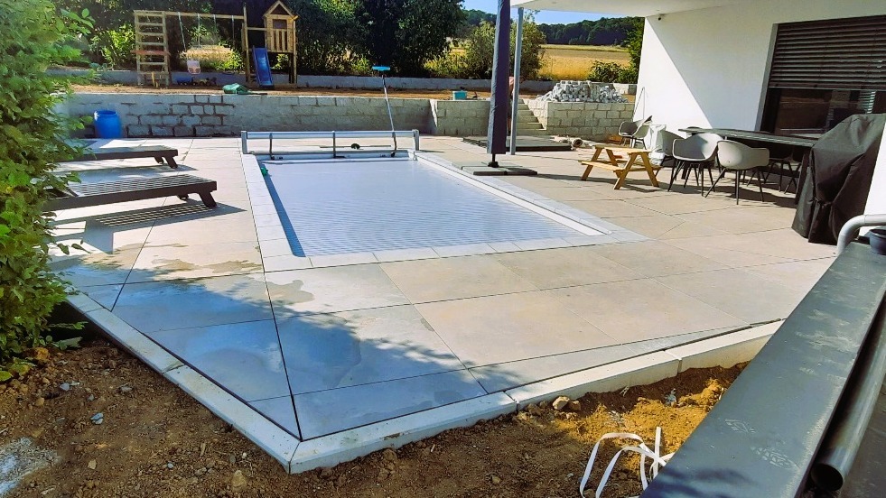 Nowoczesny taras z jasnych płyt wokół basenu z roletą, w tle meble ogrodowe i zieleń. Widoczne niedokończone prace ziemne przy obrzeżach tarasu.