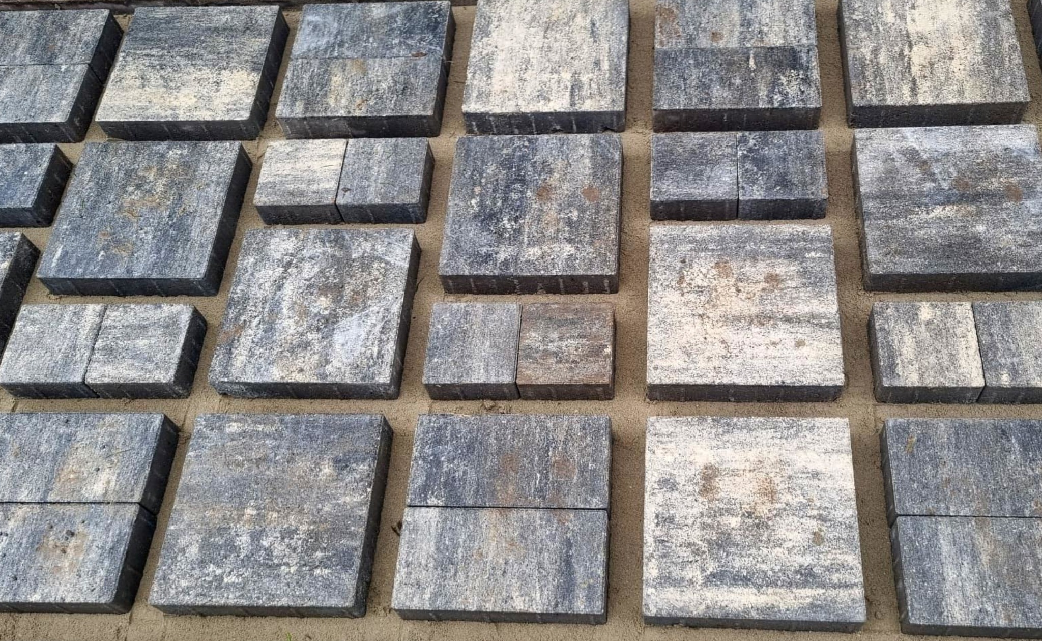 Szare, kwadratowe kostki brukowe ułożone na piasku, widoczne z góry. Kostka o zróżnicowanej fakturze i odcieniach szarości, tworząca regularny wzór.