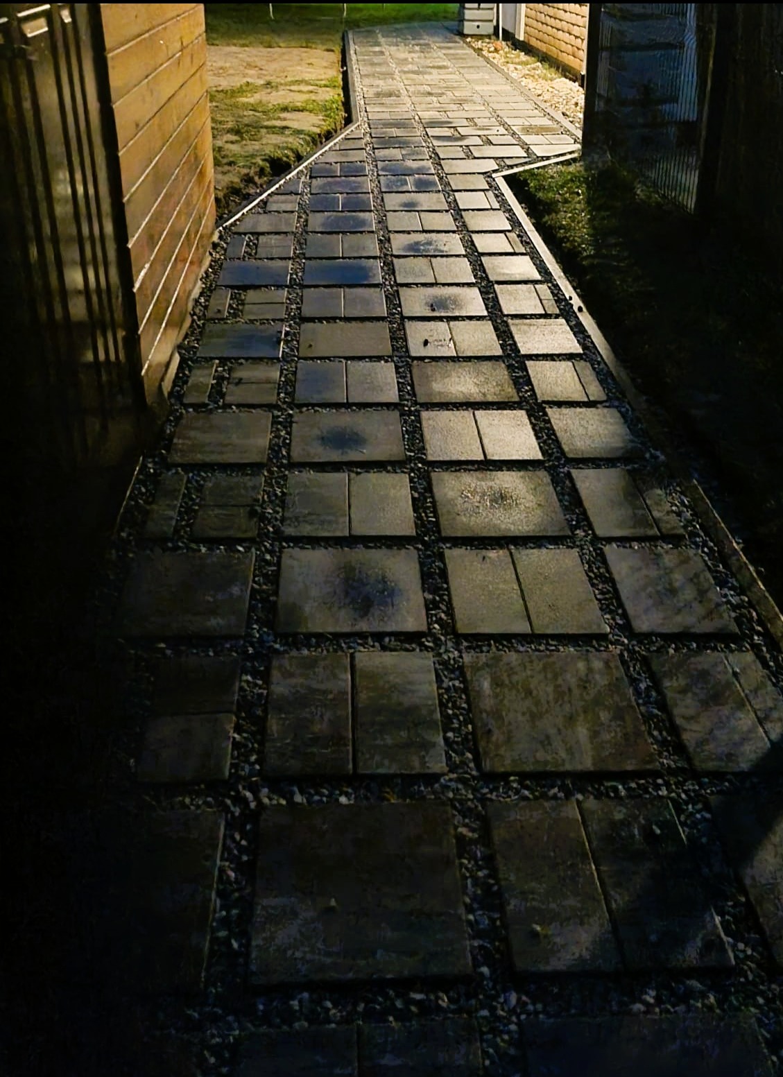 Ścieżka z kwadratowych płyt, wypełniona drobnym żwirem, prowadząca wzdłuż drewnianego ogrodzenia. Ujęcie z perspektywy, wieczorne oświetlenie.