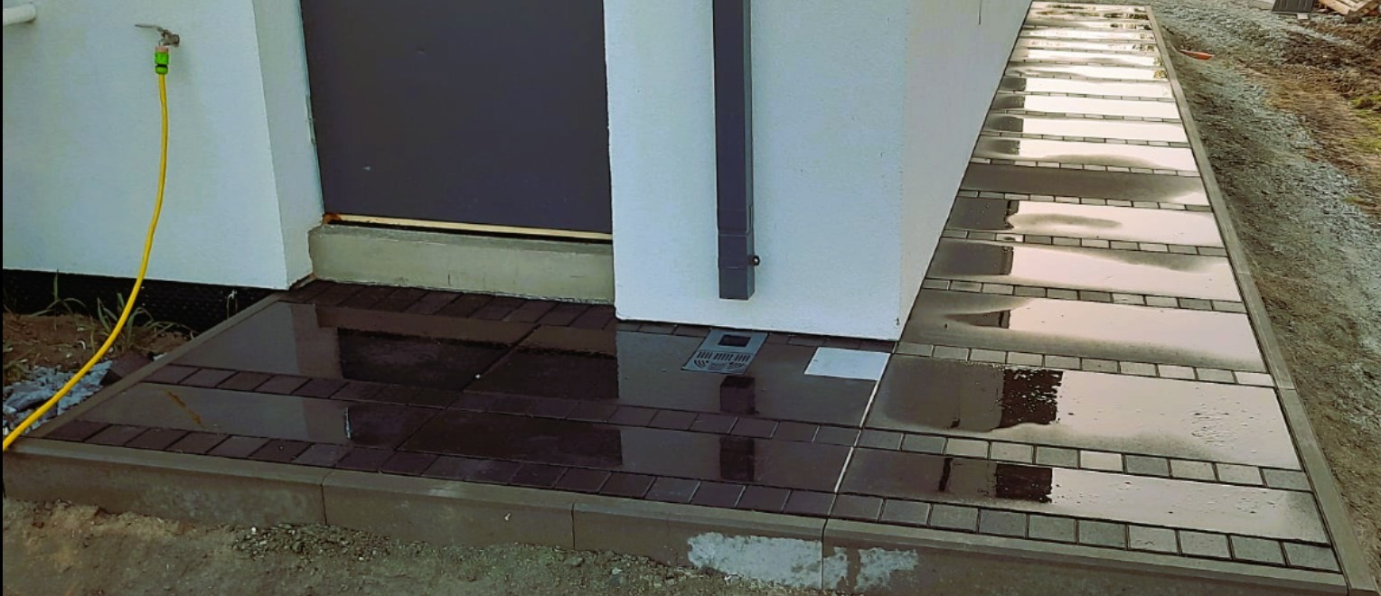 Nowoczesna nawierzchnia z ciemnej kostki przy budynku, po deszczu. Widoczny odpływ liniowy i żółty wąż ogrodowy. Minimalistyczny design.