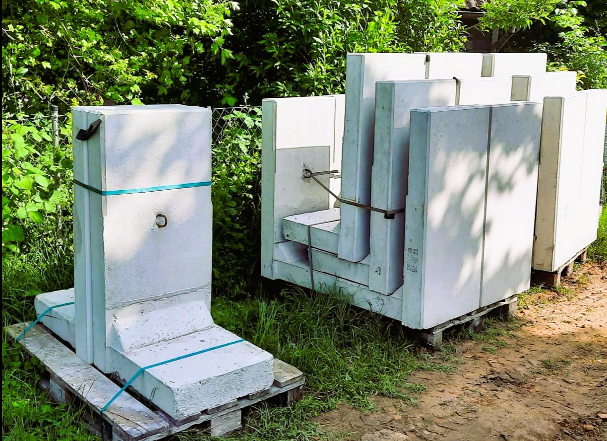 Prefabrykowane elementy betonowe na paletach, zabezpieczone taśmami, gotowe do montażu. Widoczne detale konstrukcyjne i faktura betonu. Tło: zieleń.