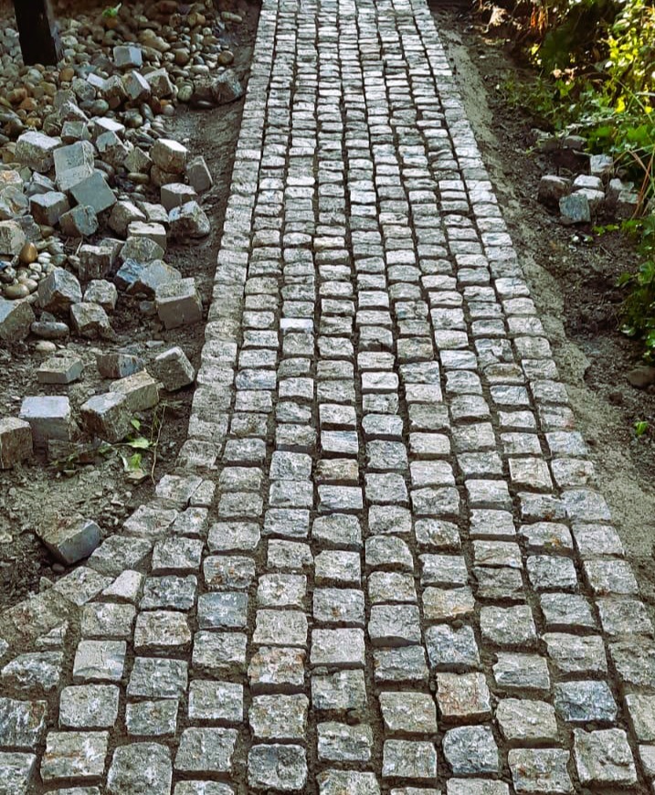 Ścieżka z kostki granitowej w ogrodzie, widok z góry. Częściowo ułożona, z boku stos kostek do ułożenia. Naturalne oświetlenie, ziemiste kolory.