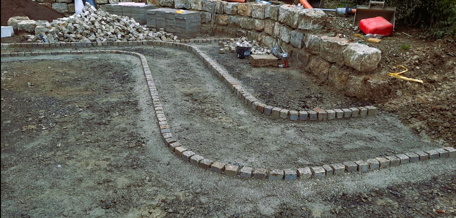 Etap prac brukarskich: obrzeża z kostki granitowej wyznaczają ścieżkę na gruncie. W tle mur z kamienia, stosy kostki i narzędzia.