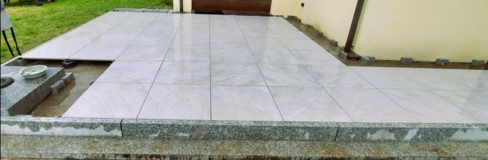 Jasnoszary taras z płytek, częściowo w trakcie układania, z granitową obwódką. Widoczne podpory pod płytami i narzędzia. Ujęcie z niskiej perspektywy.