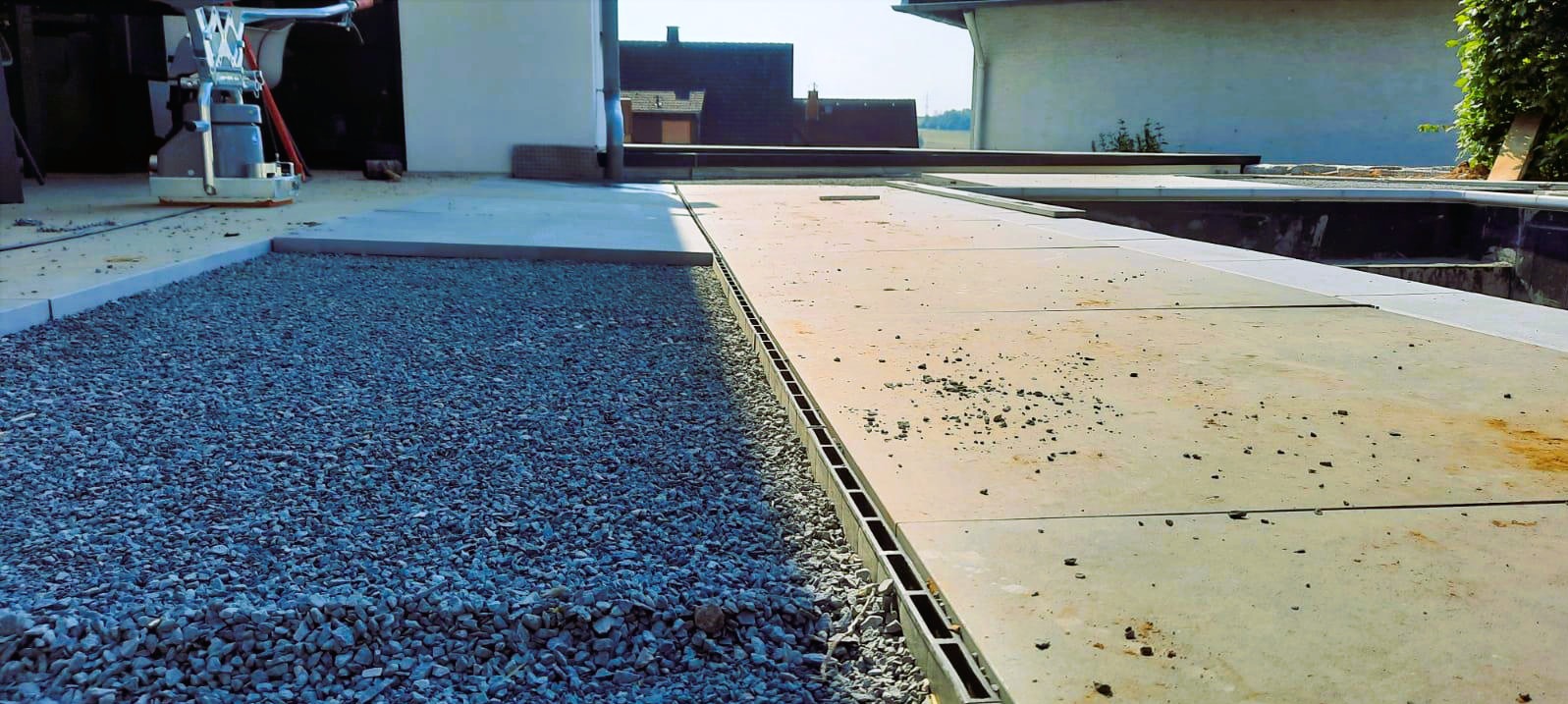 Wyrównana powierzchnia z szarego żwiru sąsiadująca z płytami tarasowymi oddzielona betonową obwódką. W tle budynek i maszyna do zagęszczania.