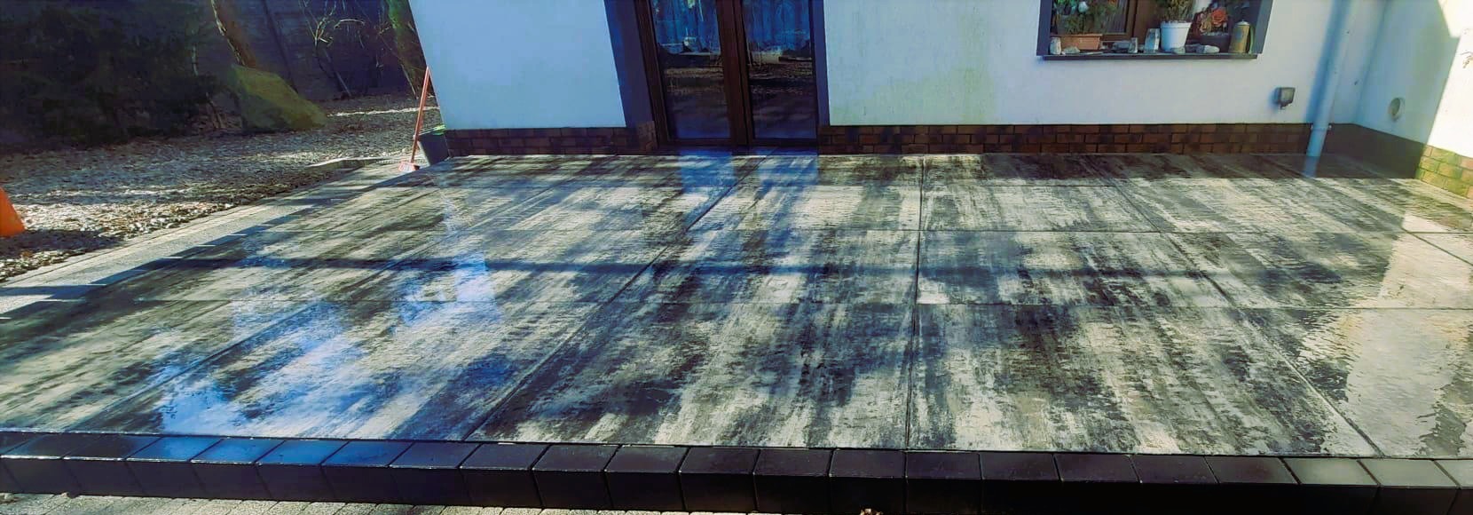 Nowoczesny taras z szarych płyt betonowych o fakturze drewna, obrzeża z ciemnej kostki. Widok na elewację domu z oknem i drzwiami tarasowymi.