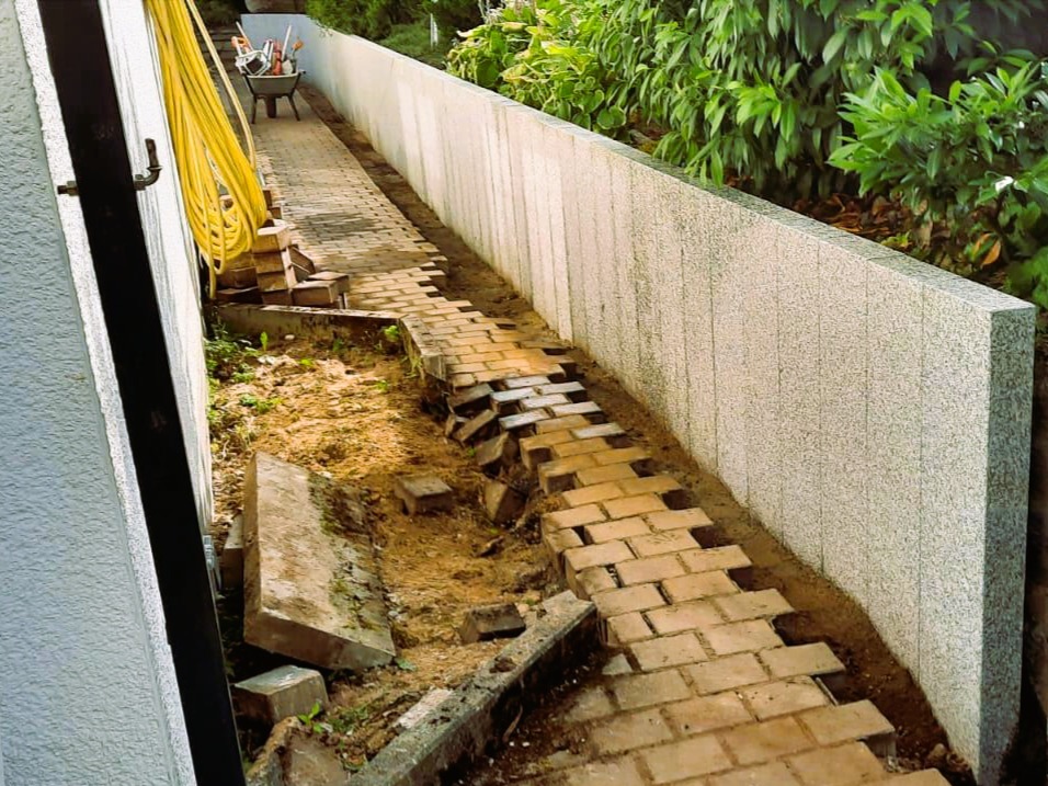 Fragment ścieżki z ceglanej kostki w trakcie układania, widoczna ziemia i narzędzia. W tle wysoki, jasny mur i zieleń.