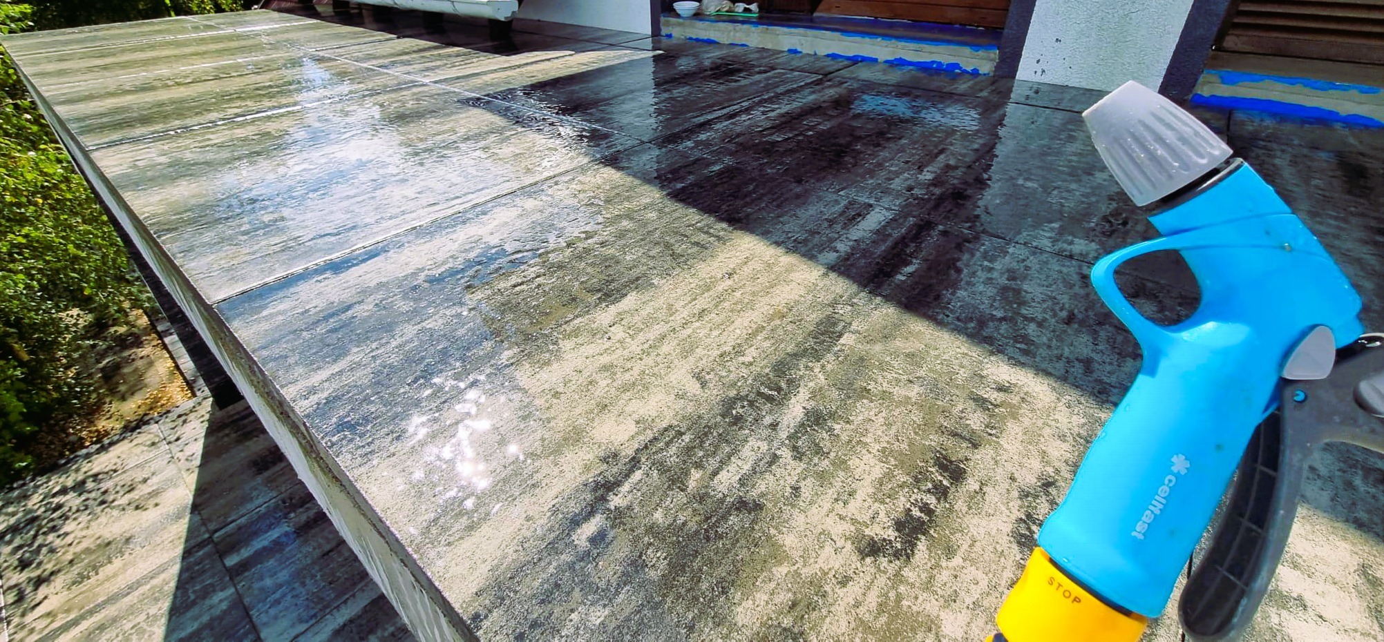 Błyszczący, mokry taras z szarych płyt po umyciu, z widocznym niebieskim pistoletem do podlewania. Widoczne refleksy światła na powierzchni.