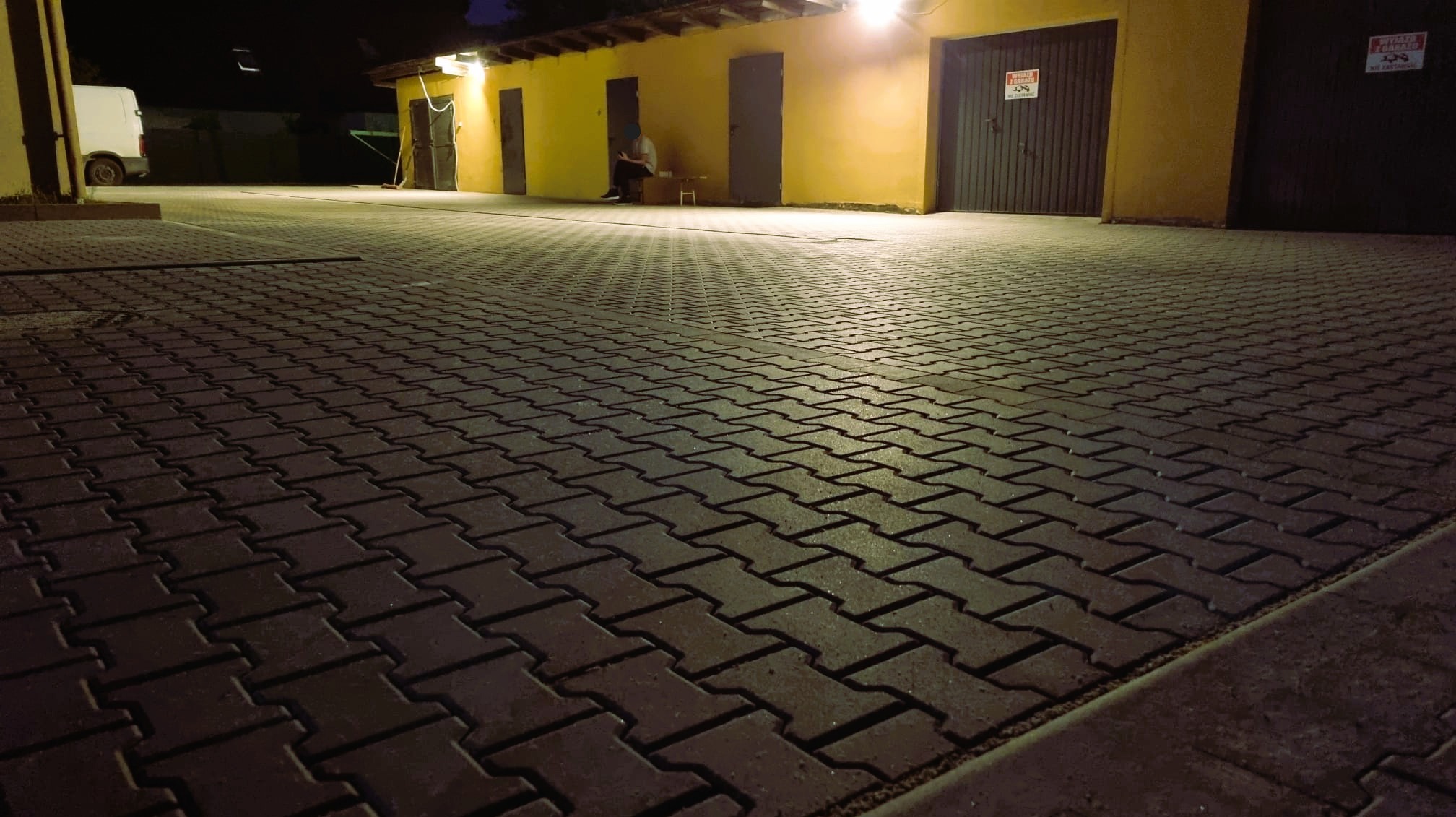 Ułożona, ciemna kostka brukowa na podjeździe, oświetlona nocą. W tle żółty budynek z bramami garażowymi i siedząca osoba. Widok z niskiej perspektywy.