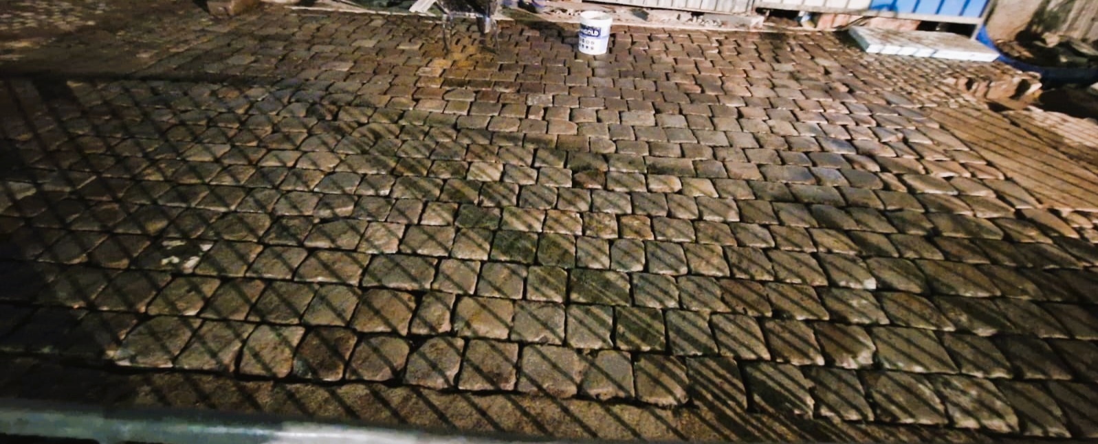 Ułożona kostka granitowa w jodełkę, widok z góry, z widocznym wiaderkiem farby w tle. Ciemne oświetlenie i cienie rzucane przez ogrodzenie.