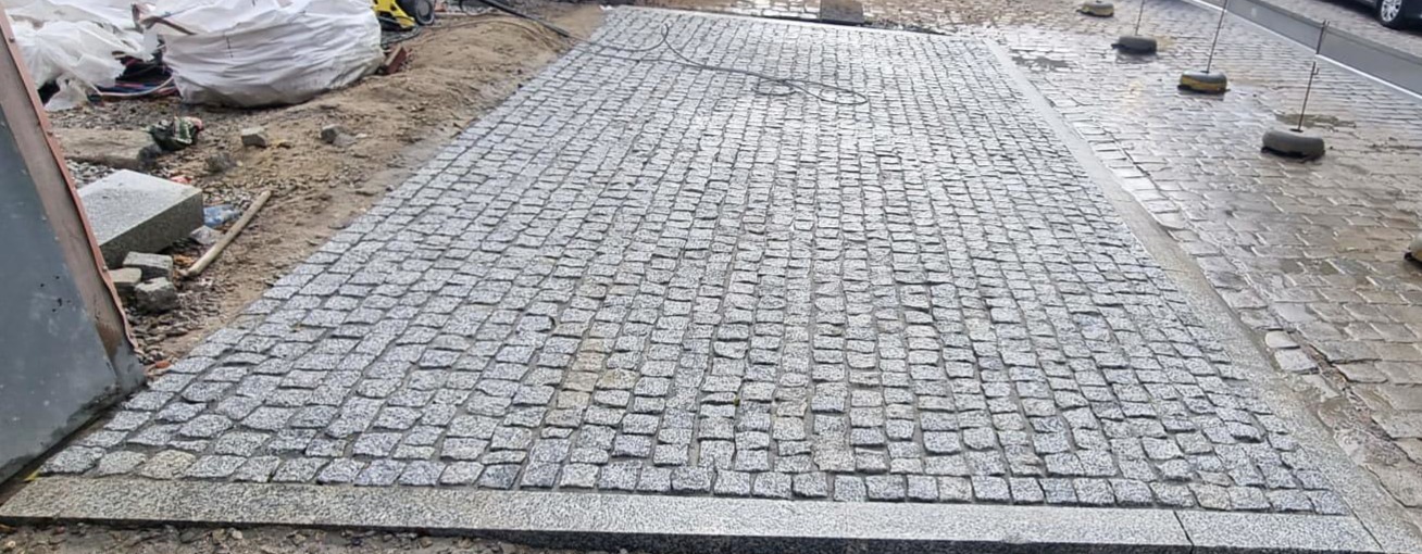 Świeżo ułożona, szara kostka granitowa tworzy równą nawierzchnię. Widoczne worki z materiałem budowlanym i sprzęt w tle. Krawężnik oddziela kostkę od jezdni.